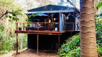 A rustic cabin with a wooden deck, surrounded by dense forest.
