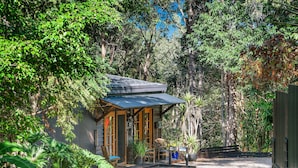 A small wooden cabin with a sloped roof, surrounded by dense forest. There is a wooden deck with a chair and a table, and a sign indicating a parking area.