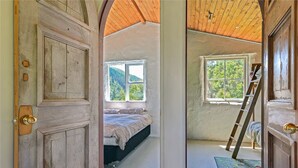 Ein rustikales Schlafzimmer mit Holztür, einem Bett, einem Fenster mit Blick auf Grünflächen und einer an der Wand angelehnten Leiter.