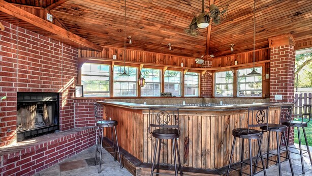 An outdoor kitchen with a brick fireplace, wooden ceiling, and a bar area with stools.