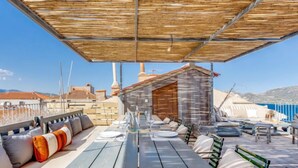 Un espace extérieur aménagé avec des tables et des chaises en bois, une partie couverte avec un toit en bois, et une vue sur la mer et les bâtiments en arrière-plan.