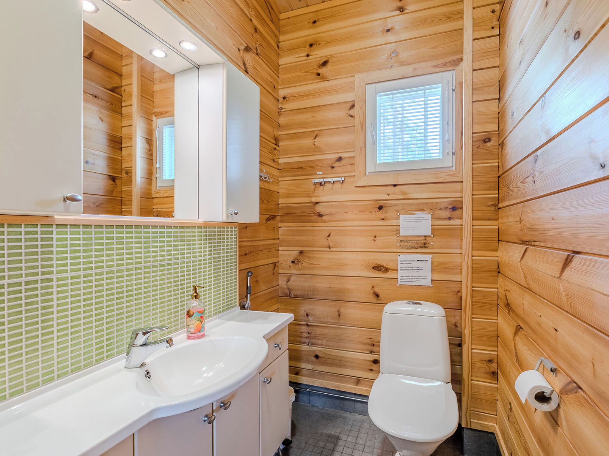 A wooden cabin bathroom with a sink, mirror, toilet, and a small window.