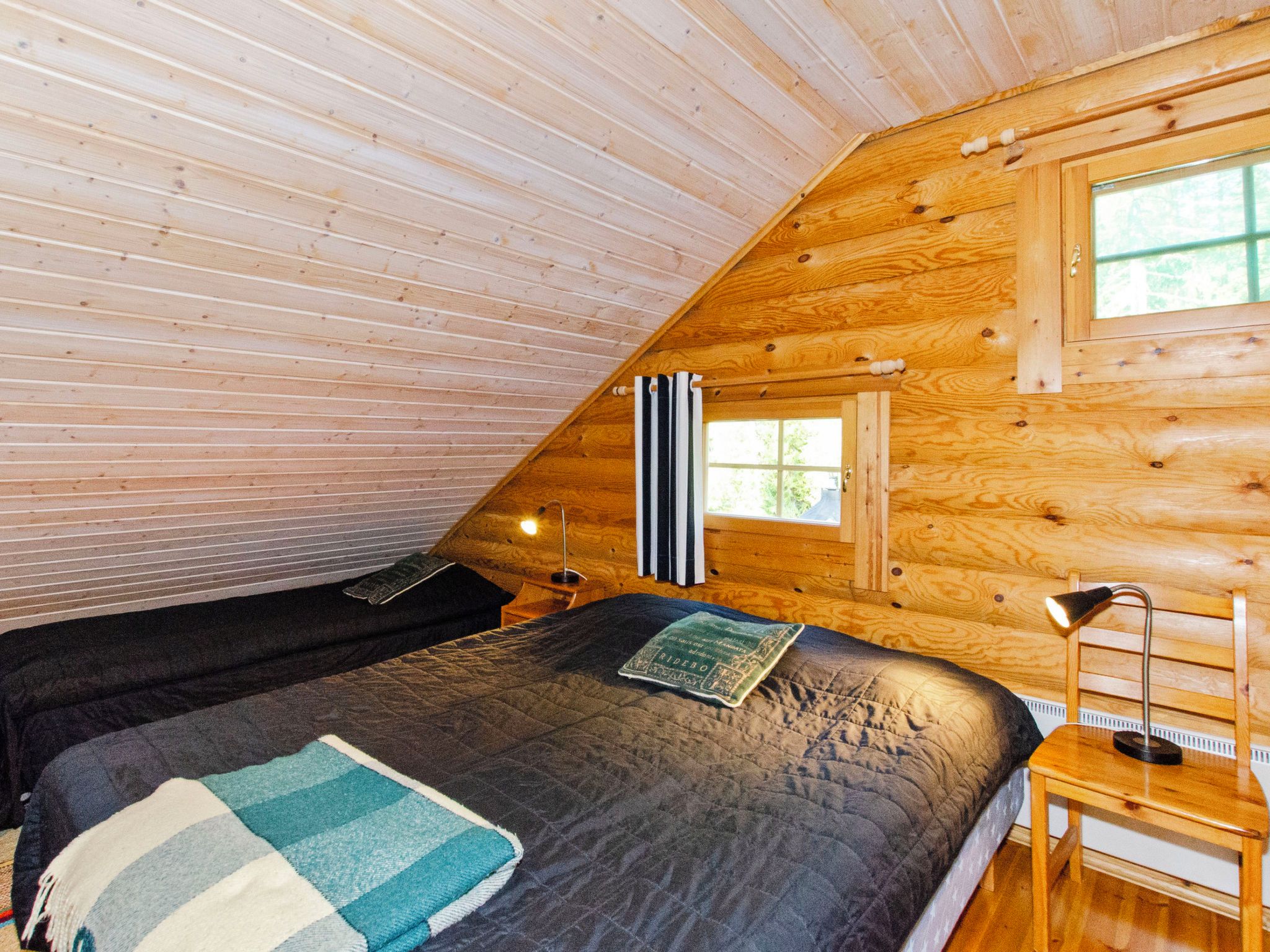 Une chambre mansardée chaleureuse, avec un plafond en pente, un lit simple recouvert d’une courtepointe, une table de chevet avec une lampe et une fenêtre avec des rideaux.