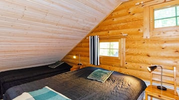 Une chambre mansardée chaleureuse, avec un plafond en pente, un lit simple recouvert d’une courtepointe, une table de chevet avec une lampe et une fenêtre avec des rideaux.