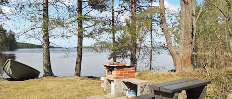 A picnic table and a brick barbecue grill near a lake with trees.