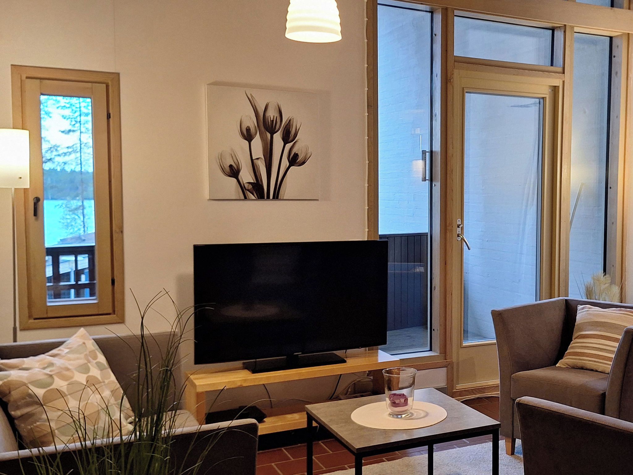 Ein Wohnzimmer mit einem Flachbildfernseher auf einem Standfuß, einem Couchtisch, einer Couch und einem Sessel. Ein Fenster bietet Blick auf Bäume und eine Tür mit Glasverzierungen.