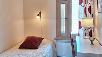 Ein Schlafzimmer mit einem Einzelbett, einem Nachttisch, einer Lampe, einem Fenster mit Jalousien und einer Wand mit einem gemusterten Vorhang.
