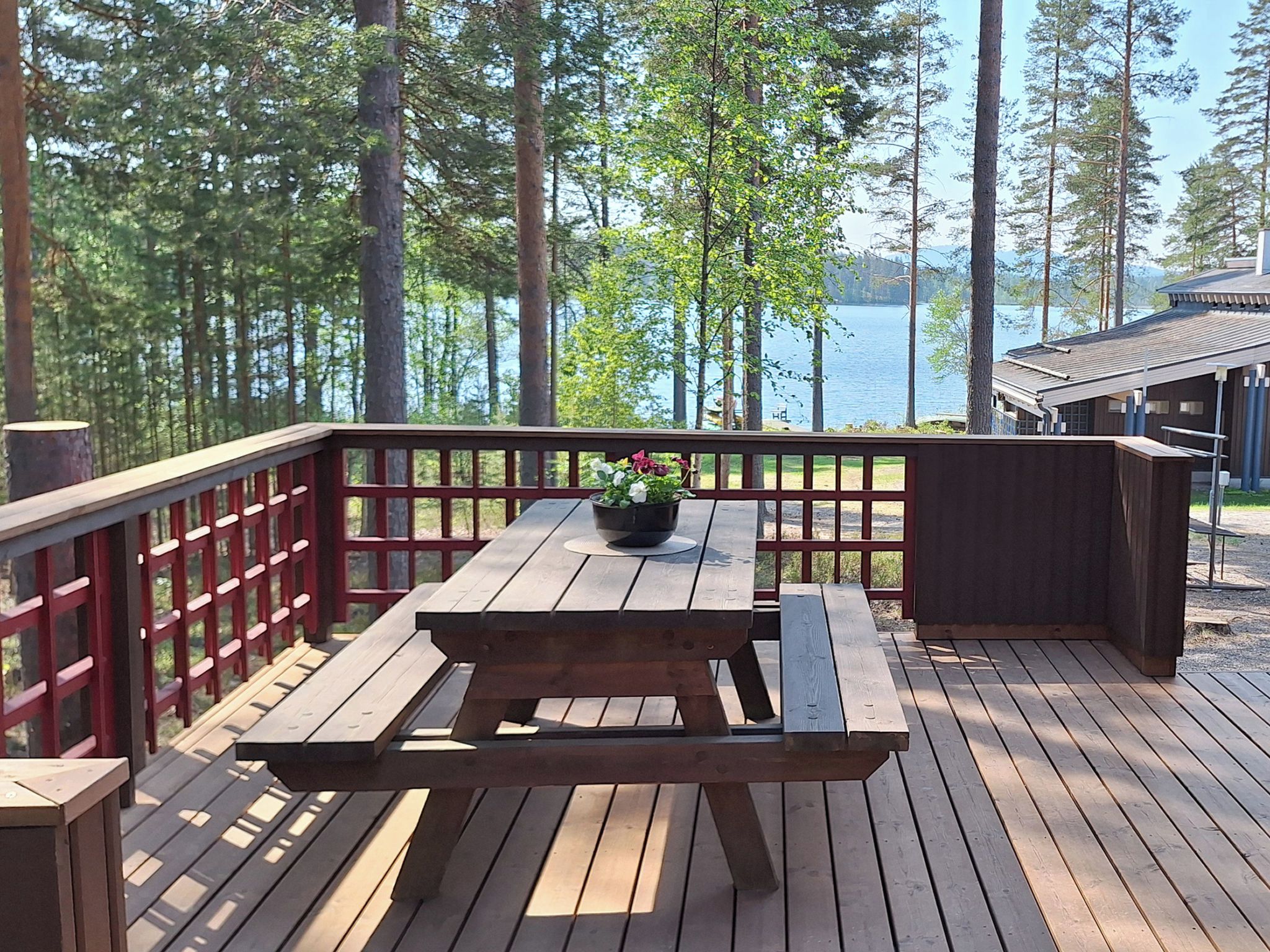 Una terraza de madera con una mesa de picnic y bancos, con vistas a un lago, árboles y una cabaña al fondo.