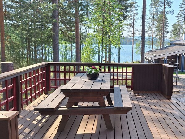 Een houten terras met een picknicktafel en banken, met uitzicht op een meer met bomen en een vakantiehuis op de achtergrond.