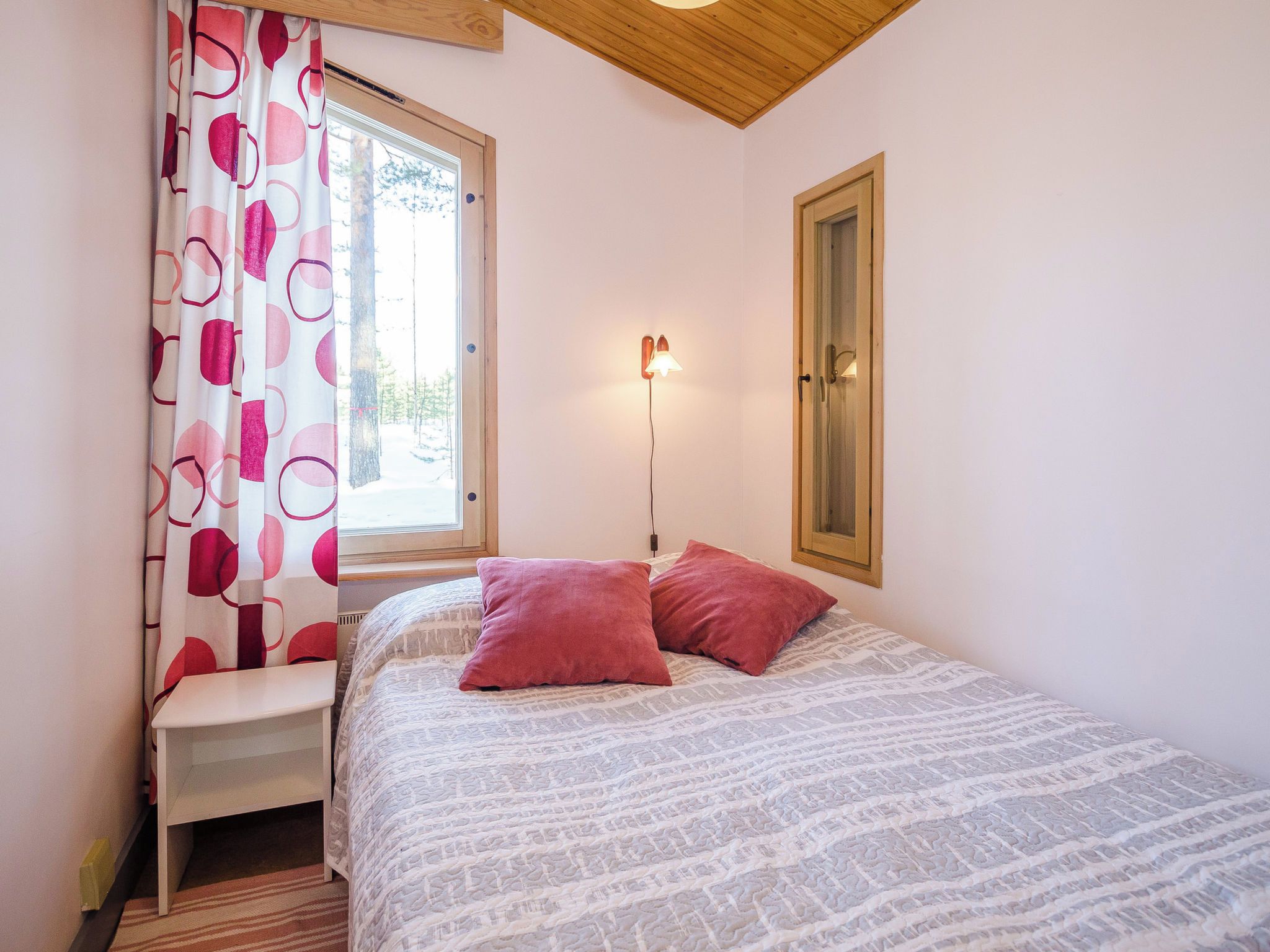 A bedroom with a bed, pillows, a nightstand, a window with curtains, a wall-mounted lamp, and a door.