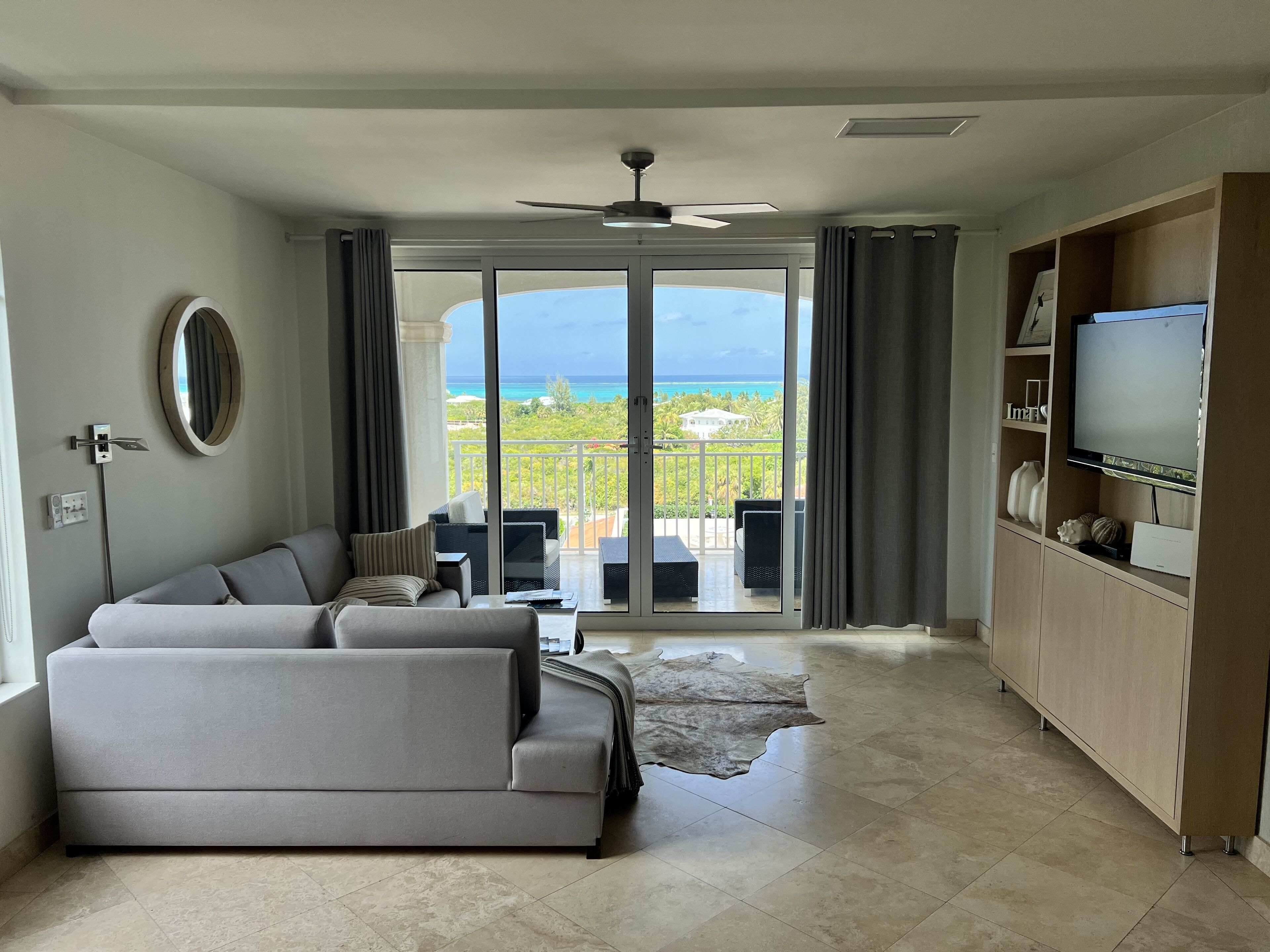 Ein modernes Wohnzimmer mit grauem Sofa, einem Flachbildfernseher und einem Balkon mit Meerblick.