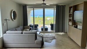 A modern living room with a grey sofa, a flat-screen TV, and a balcony with a view of the ocean.