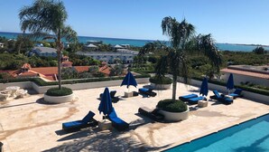 A rooftop pool area with lounge chairs and umbrellas, overlooking a coastal landscape with buildings and a clear sky.