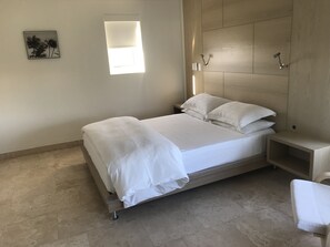 Een opgemaakt bed met witte lakens, een nachtkastje met een lamp en een klein plankje.