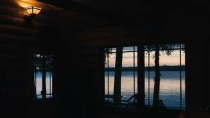Una habitación tenuemente iluminada con un ventanal grande que da a una masa de agua tranquila, techo de madera y una sola mesa con sillas.