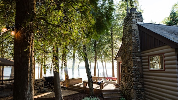 Una cabaña con chimenea de piedra, terraza de madera y vista al lago.