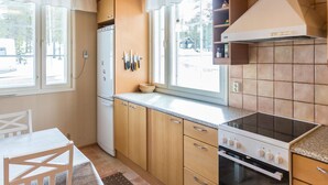 A compact kitchen with wooden cabinets, a white refrigerator, a stove, and a window with curtains.