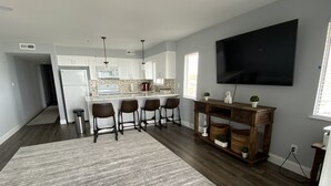 A modern kitchen with white cabinets, a central island, and bar stools. There is a large flat-screen TV mounted on the wall. - Wake up to the sunrise over the Atlantic! Beautiful ocean views from balcony  (North Myrtle Beach)