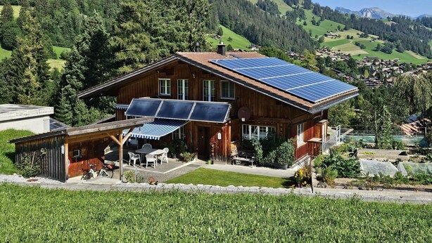 A wooden house with solar panels on the roof, a patio with outdoor furniture, and a view of a green valley and mountains in the background. - Burkhalter by Interhome (Zweisimmen)