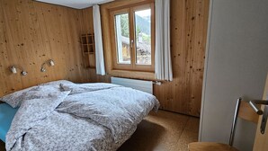 A wooden-paneled room with a bed, a window with curtains, a radiator, and a wooden chair. - Burkhalter by Interhome (Zweisimmen)