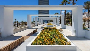 A modern architectural structure with a central garden of yellow and orange flowers, white walls, and a clear view of the sea. - Reina Sofia II by Interhome (Estepona)