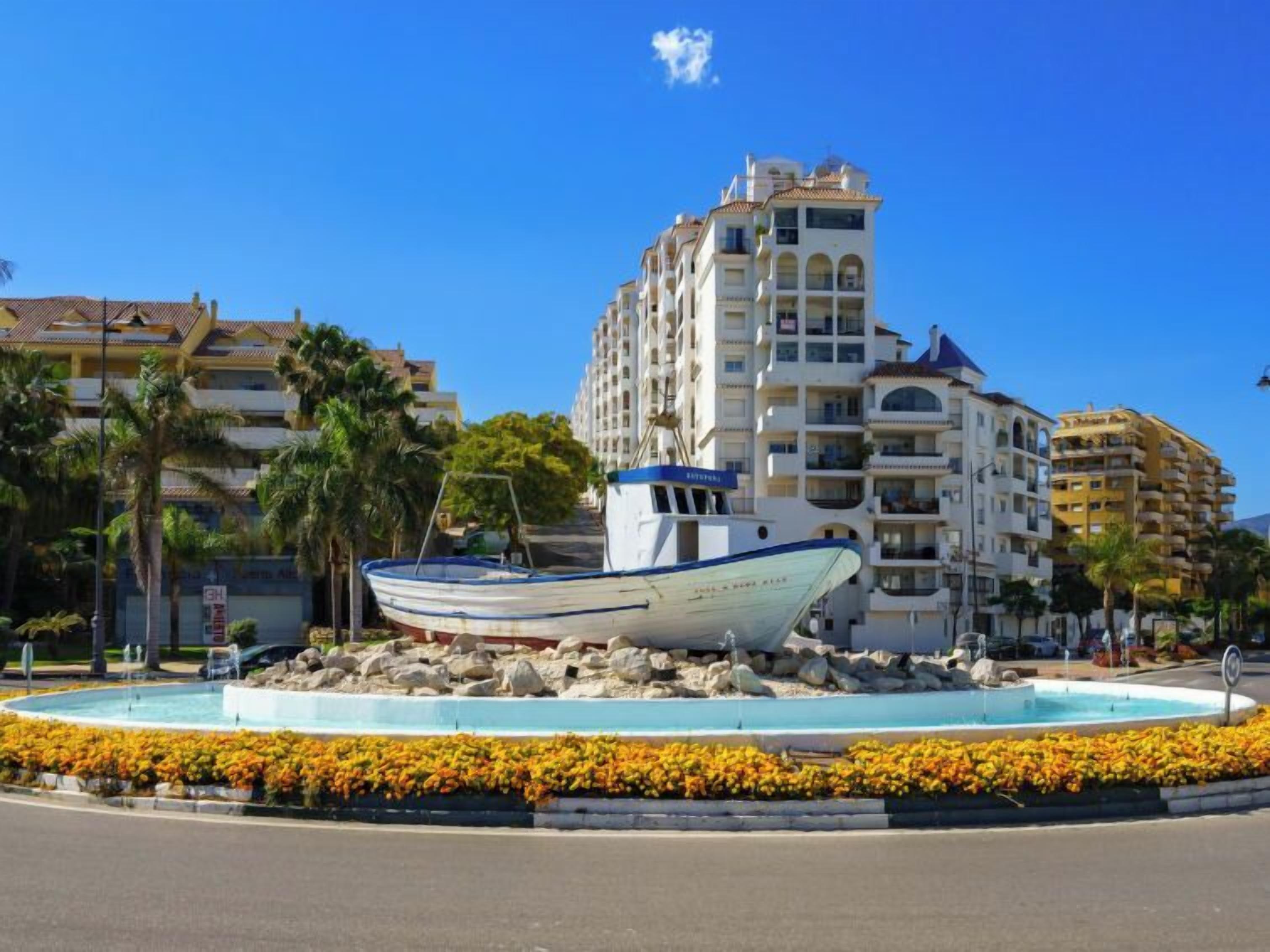 Una escultura de barco en una fuente circular rodeada de flores coloridas, con edificios modernos al fondo.