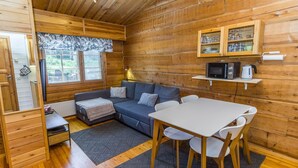 Un intérieur de chalet chaleureux avec un revêtement mural en bois, un canapé, une table à manger avec des chaises, un coin cuisine équipé d’un four à micro-ondes et d’une bouilloire, et une fenêtre avec des stores.