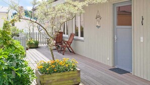 Eine Holzdielen-Terrasse mit weißem Zaun, einem kleinen Tisch mit Stühlen und einem Blumentopf mit gelben Blüten.