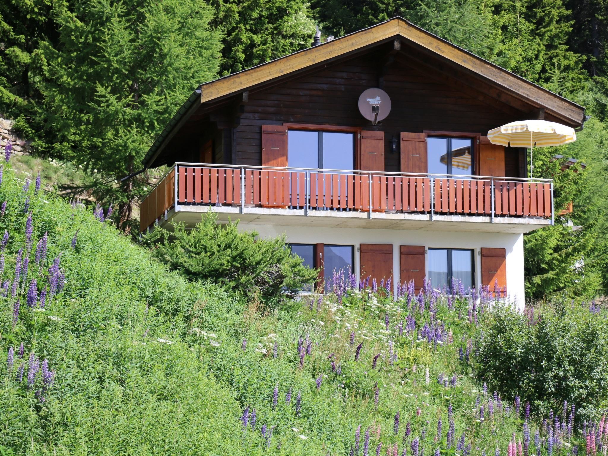 Un chalet en bois à deux étages, doté d’un balcon, entouré d’une végétation luxuriante et de fleurs violettes.