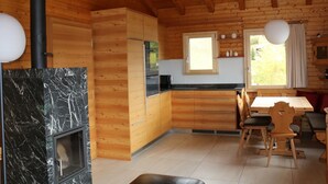 A wooden cabin with a kitchen featuring a marble fireplace, wooden cabinets, and a dining area with chairs and a table. - Brunnquell by Interhome (Bettmeralp)