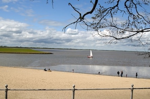 Une plage de sable fin, une étendue d’eau calme, un voilier et des personnes sur le rivage.