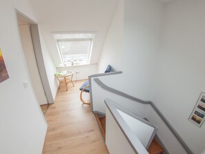 Un couloir étroit doté d’un plancher en bois, une petite table avec une chaise, une fenêtre avec des stores et un mur blanc.