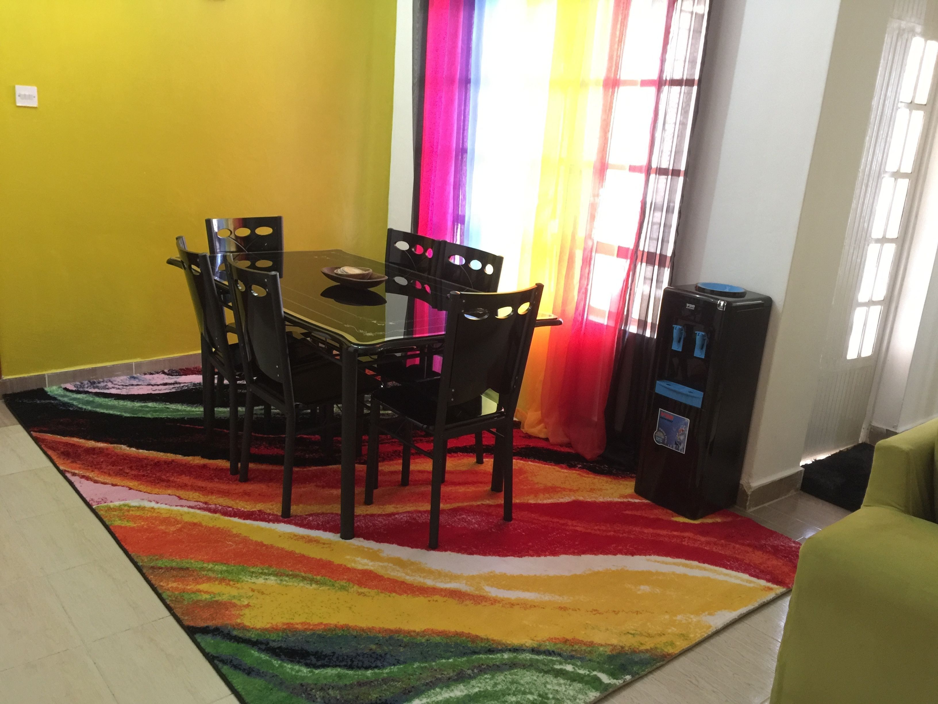 A dining area with a table and chairs, a colorful rug, a refrigerator, and a window with curtains.