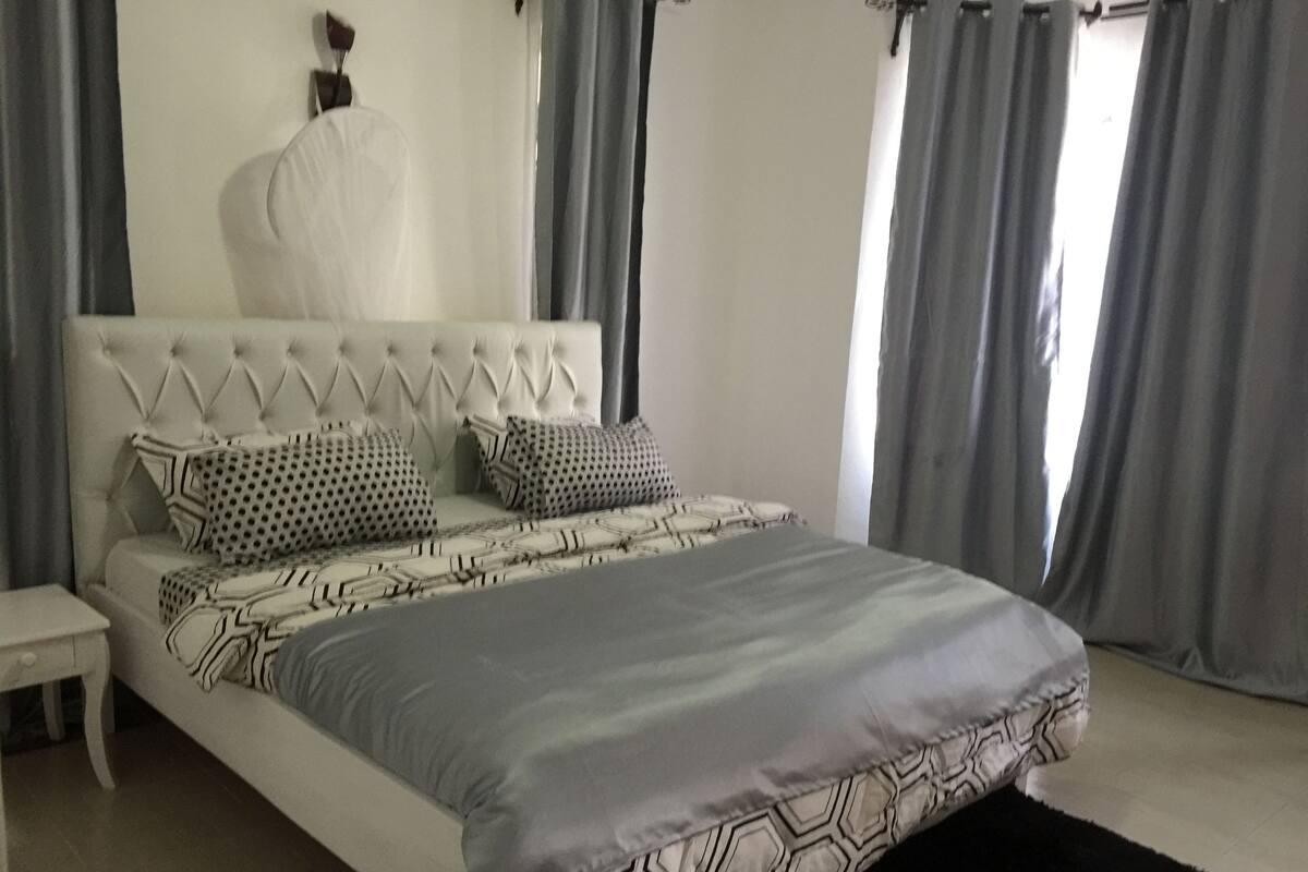 A bedroom with a tufted headboard, patterned bedding, a bedside table, and grey curtains.