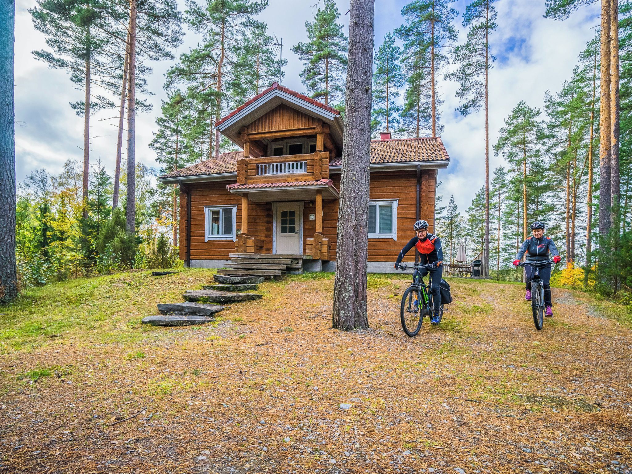 A two-story wooden cabin with a porch, surrounded by a forest.