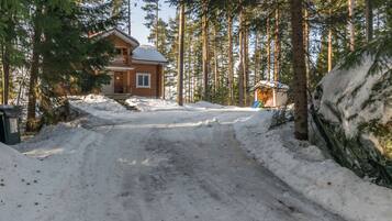 Une cabane en bois, dont l’allée est recouverte de neige, entourée de grands pins.