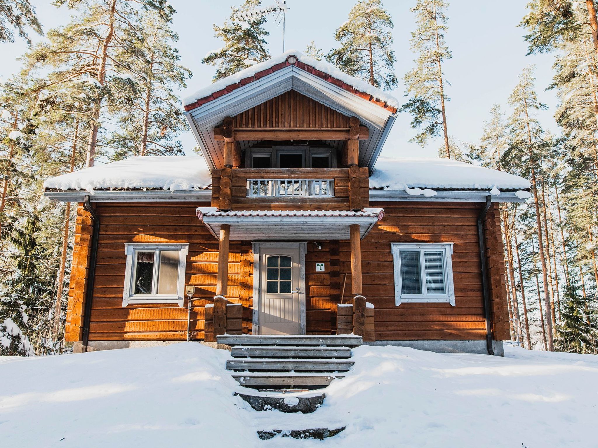 Een houten chalet met een veranda, schoorsteen en een sneeuwbedekte dak, omringd door hoge dennenbomen.