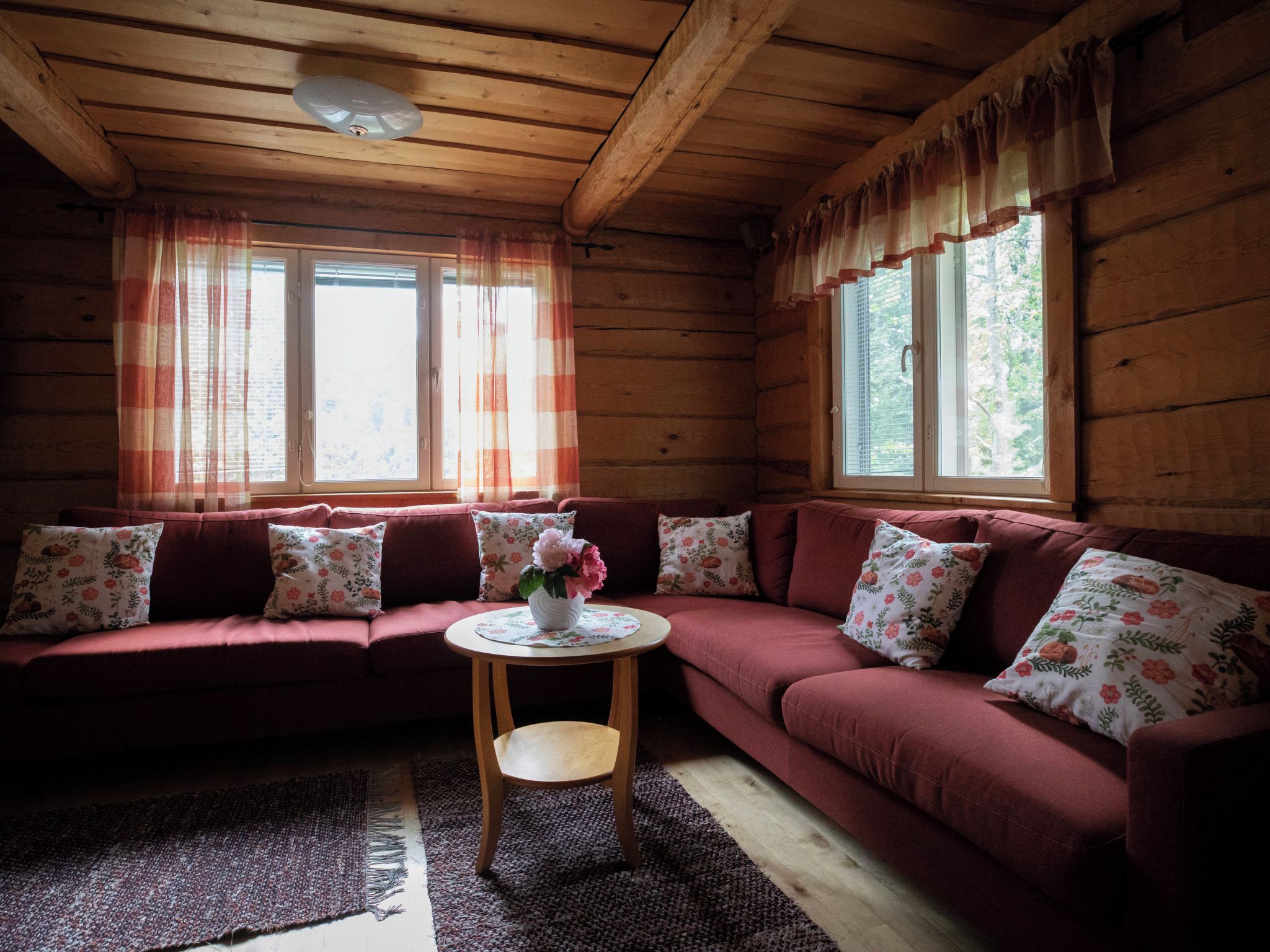 A cozy cabin interior with a wooden ceiling, a red sofa with floral cushions, a small round table with a vase, and large windows with curtains.