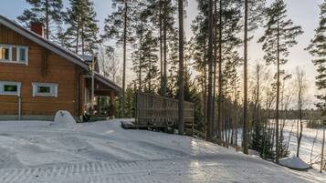 A wooden cabin with a porch, surrounded by a snowy landscape and tall pine trees.