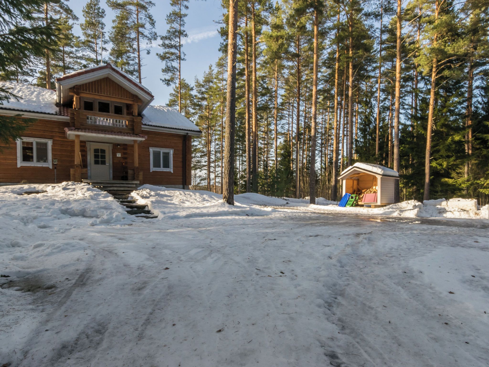 Puinen mökki, jossa on kuisti ja pieni varasto, ja sen ympärillä on lunta sekä korkeita mäntyjä.