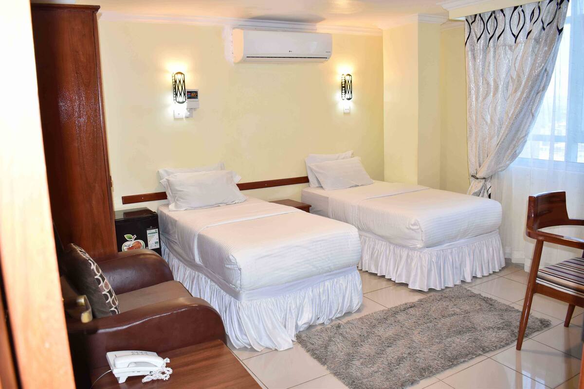 A hotel room with two beds, a sofa, a chair, an air conditioner, and a window with curtains.