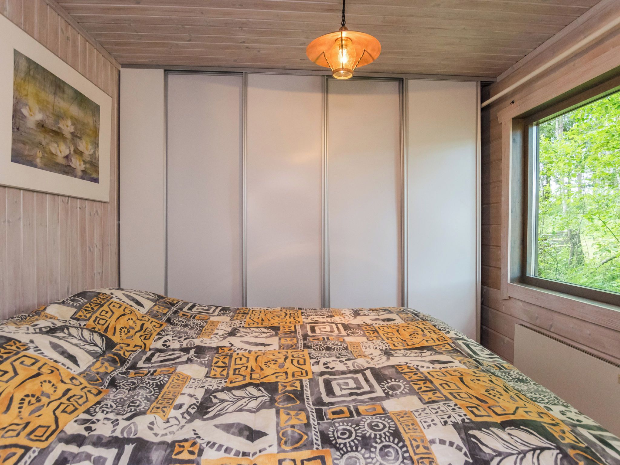 A bedroom with a patterned bedspread, a painting on the wall, a window with a view of greenery, and a hanging light fixture.