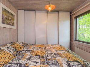 A bedroom with a patterned bedspread, a painting on the wall, a window with a view of greenery, and a hanging light fixture.