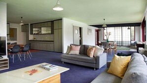 A spacious living area with a grey sofa, a coffee table, and a dining table with chairs. There is a kitchen in the background with cabinets and a window. - Stylish, Central, 3-Bedroom, 2-Bath Apartment in St Kilda with pool & trampoline (St Kilda)