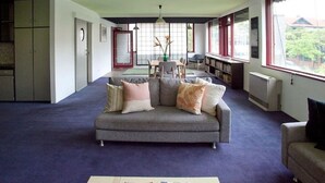 A living room with a grey sofa, a coffee table, and a bookshelf. There is a dining area with a table and chairs, and a window with a view of trees. - Stylish, Central, 3-Bedroom, 2-Bath Apartment in St Kilda with pool & trampoline (St Kilda)