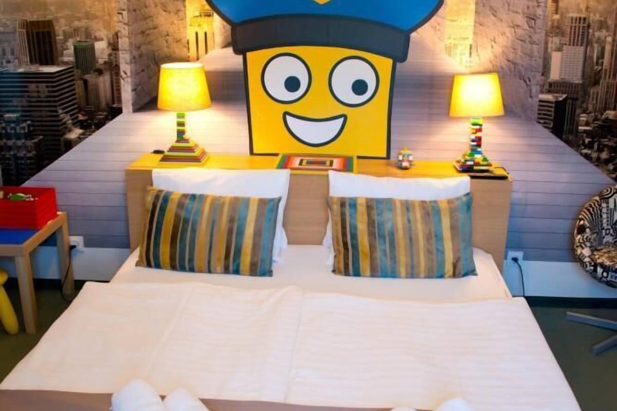 Habitación de hotel con una cama grande, dos almohadas, dos lámparas y un adorno de pared tamaño grande con un personaje de dibujos animados usando un gorro de policía.