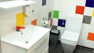Un baño con un lavamanos blanco, un inodoro y un cubo de basura. Las paredes están cubiertas de azulejos cuadrados multicolores.