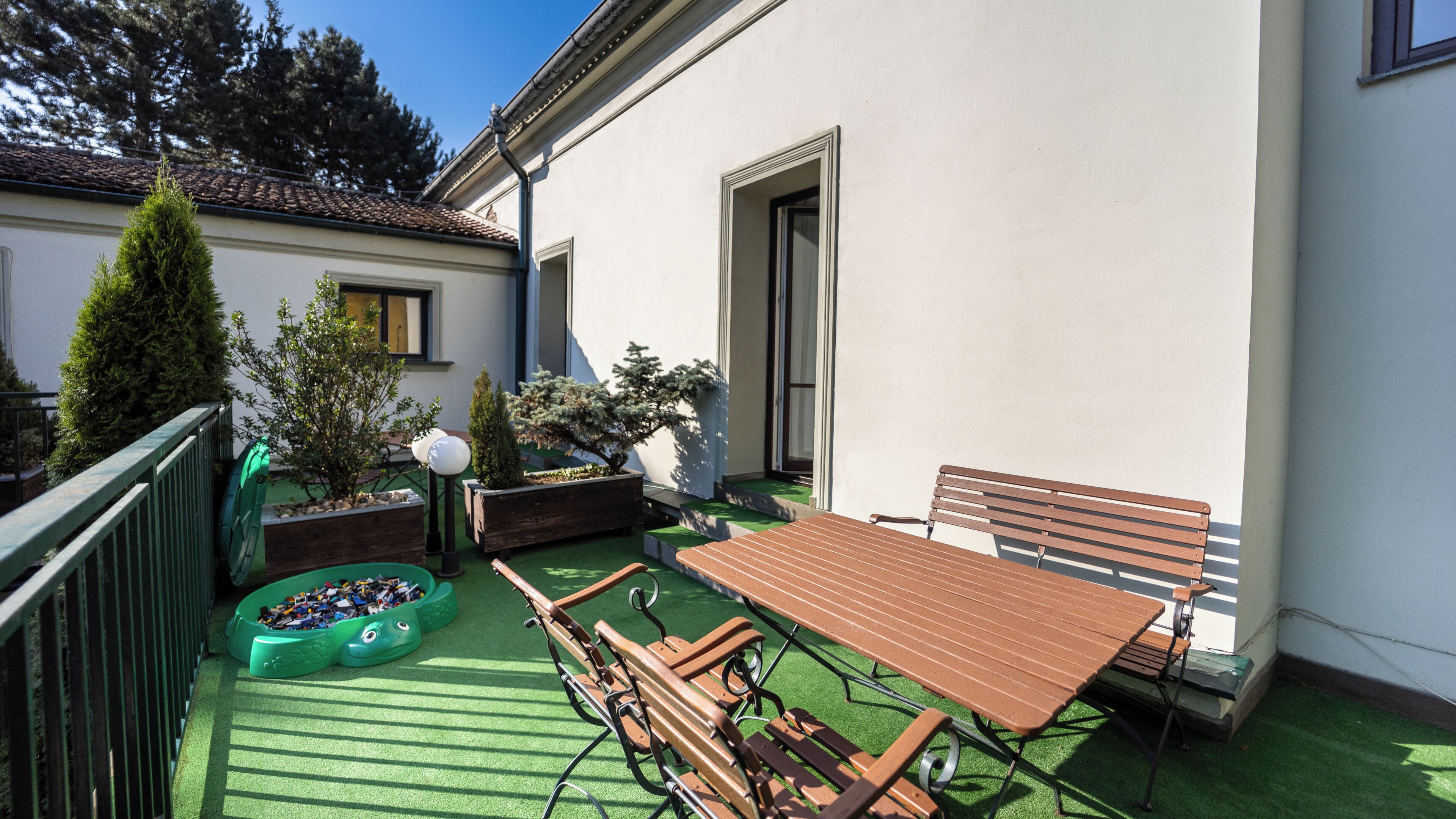 Een terras met een houten tafel en bankjes, omringd door potplanten en kunstgras.