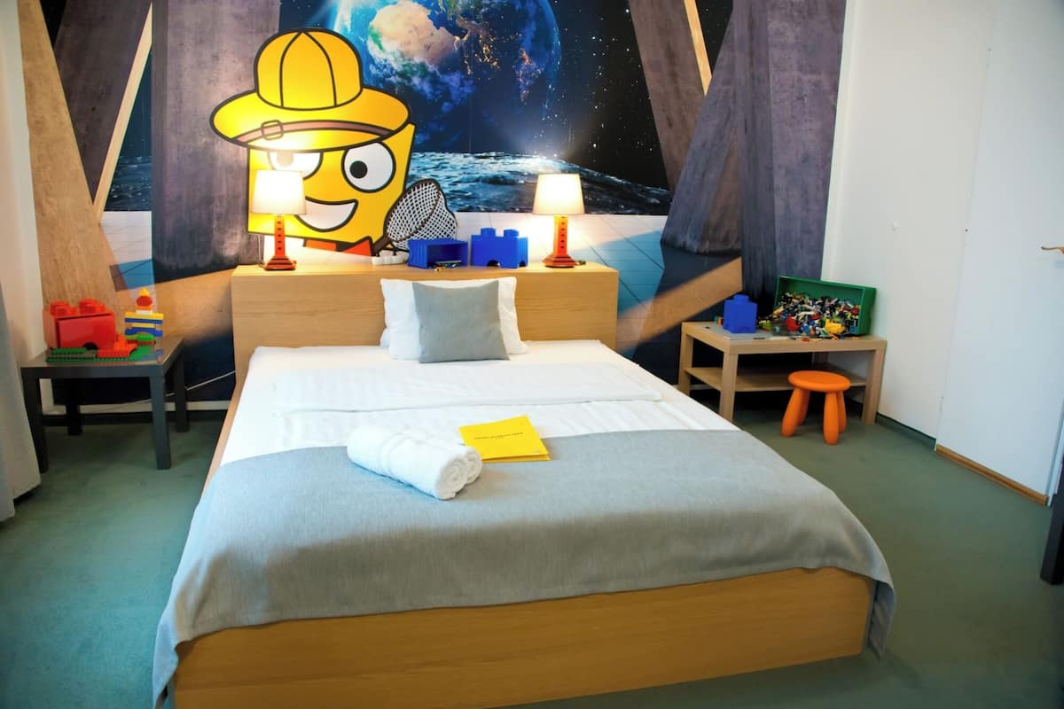 Habitación de hotel con cama, mesita de noche, escritorio y silla. Hay un cartel grande en la pared y una ventana con vista al cielo nocturno.