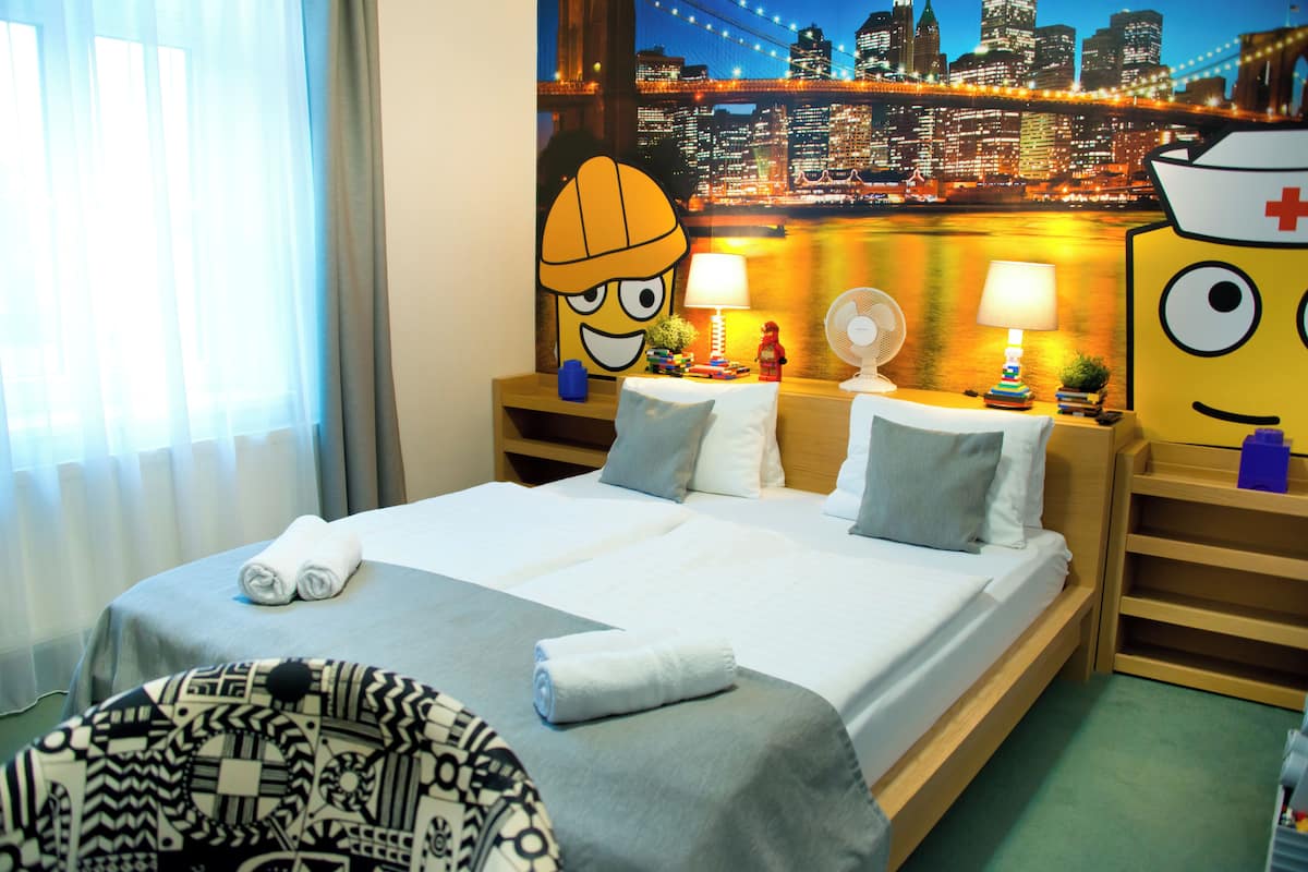 Habitación de hotel con cama, dos mesitas de noche, lámpara, ventilador y un gran ventanal con cortinas.